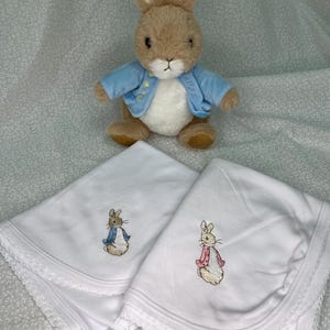 Może przedstawiać: Dwa białe kocyki dla niemowląt z haftowanymi wzorami Królika Piotrusia. Jeden kocyk przedstawia Królika Piotrusia w niebieskiej kurtce, drugi przedstawia Królika Piotrusia w różowej kurtce. Oba kocyki mają białą koronkową lamówkę.