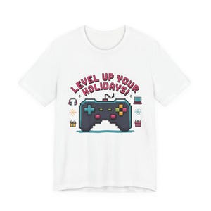 Puede incluir: Camiseta blanca con un diseño de mando de videojuego pixelado y el texto "LEVEL UP YOUR HOLIDAYS!". El mando tiene botones azules, verdes, amarillos y rojos. Incluye gráficos de auriculares, portátil, copos de nieve y cajas de regalo.