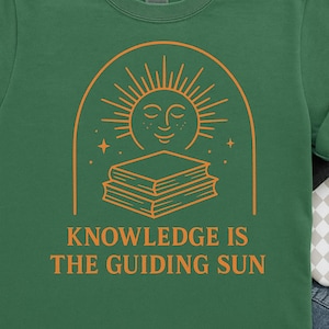 May include: Green t-shirt with an orange graphic of a smiling sun above a stack of books, within an arch. The text "KNOWLEDGE IS THE GUIDING SUN" is below the books. The shirt has a crew neck and short sleeves.