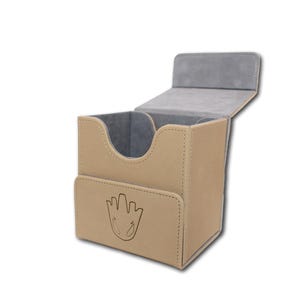 May include: Tan card storage box with a hinged lid, open to reveal a grey interior. The front features a flap with a stylized hand print design. The box is designed to hold cards.