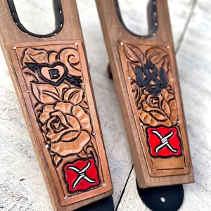 Könnte beinhalten: Paar Holz-Stiefelknechte mit Lederakzenten. Das Leder zeigt geschnitzte Blumenmuster, ein Herz mit dem Buchstaben "B" und ein rot-weißes Emblem. Die Stiefelknechte haben eine dunkelbraune Oberfläche und schwarze Gummiböden.