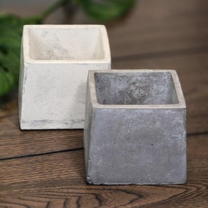 May include: Two square concrete planters, one light gray and one darker gray, sit side-by-side. The planters have a rough, textured surface and a simple, modern design. They are suitable for small plants or succulents.