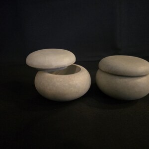 May include: Two small, round, stone-like containers with lids. One lid is slightly ajar, revealing the interior. The containers are a neutral, light gray color and sit on a dark surface. The overall aesthetic is minimalist and natural.