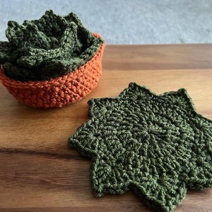 May include: A set of dark green crocheted coasters shaped like succulent leaves, with one coaster lying flat and the others stacked inside a small, round, orange crocheted bowl that mimics the appearance of a terracotta plant pot.