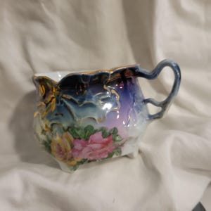 May include: A small, ornate porcelain creamer with a floral design. The creamer features a blue and purple gradient with gold trim and a delicate handle. Pink and yellow roses are painted on the side, adding a touch of elegance.