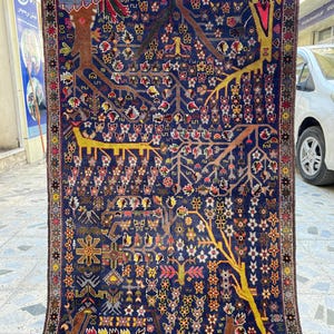 May include: A rectangular, hand-woven rug with a dark blue background and intricate patterns. The design features stylized trees in yellow and brown, with colorful floral and geometric motifs. The rug has a fringed border and a detailed border.