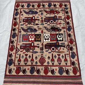 Op de afbeelding: Een rechthoekig beige tapijt met een herhaald patroon van helikopters, tanks en granaten in rood, blauw en bruin. Het tapijt heeft een gefranjerde rand en een rand met geometrische ontwerpen.