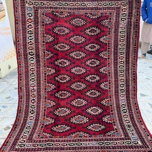 May include: A rectangular, hand-knotted rug with a deep red field and intricate geometric patterns. The rug features a central diamond motif, bordered by detailed designs in shades of blue, cream, and brown. The edges have a fringed finish.