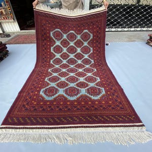 Könnte beinhalten: Ein rechteckiger, handgewebter Teppich mit einem hellblauen Mittelfeld mit einem sich wiederholenden Muster aus roten und orangefarbenen geometrischen Medaillons. Der Rand ist tiefrot mit komplizierten Designs, und der Teppich hat einen weißen Fransen.