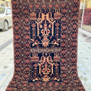 Könnte beinhalten: Ein rechteckiger, handgewebter Teppich mit einem zentralen geometrischen Design in Marineblau, Beige und Rot. Der Teppich weist komplizierte Muster und einen Fransenrand auf. Der Teppich wird hochgehalten und zeigt seine detaillierte Handwerkskunst und seinen traditionellen Stil.