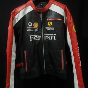 Könnte beinhalten: Schwarze, rote und weiße Lederjacke mit Ferrari-Logo und Sponsorenlogos. Die Jacke hat einen Reißverschluss vorne, rote Akzente an Schultern und Ärmeln sowie weiße Streifen. Das Ferrari-Logo befindet sich auf der Brust.