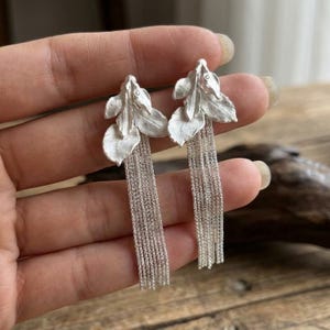 May include: A pair of silver-toned earrings featuring a matte leaf-shaped top section with a textured surface. Attached to the base of each leaf are multiple thin, dangling silver chains that create a long, fringe-like effect hanging downwards.