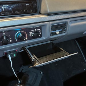 May include: Interior view of a car dashboard with a Sony stereo, climate controls, and an open storage compartment. A black gear shift knob with a white shift pattern is visible. A USB charger is plugged in.