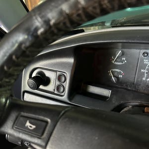 May include: Close-up of a car's dashboard and steering wheel. The black steering wheel is worn and textured. The dashboard features gauges, buttons, and a light switch. The image captures the interior of a vehicle.