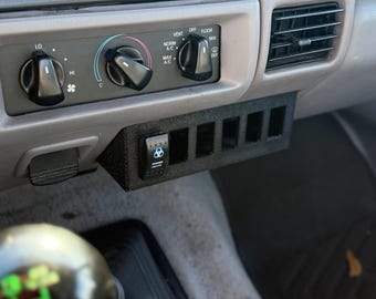 May include: A black, rectangular switch panel with six openings, mounted below a vehicle's dashboard controls. The panel includes a switch labeled "BONNER LIGHTS" and is positioned near the air conditioning and vent controls.