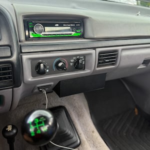 May include: Interior view of a vehicle's dashboard, featuring a JVC stereo system with green accents, climate control knobs, and a black storage compartment. A gear shift knob with green markings is visible, along with a smaller black gear shift.
