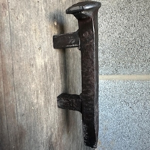 May include: A rustic, dark metal door handle, likely iron, mounted on a weathered wooden door. The handle has a rectangular shape with a flat top and two horizontal extensions. The door is made of gray wood.