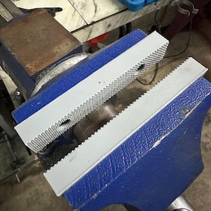 May include: A blue and gray metal vise with a textured surface. The vise has a blue base and a gray top with a serrated edge. The vise is open, ready to hold an object. Tools are visible in the background.