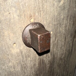 May include: A dark brown, weathered metal square-headed bolt or plug with a circular flange base. The metal surface shows signs of oxidation and wear. The text 1206 is embossed on the left side of the flange, and the text IX6B is embossed on the top.