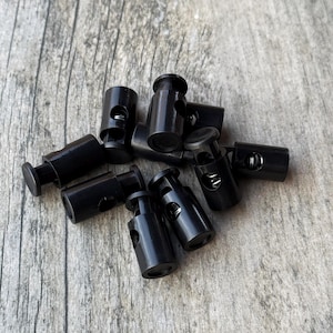 May include: A group of black plastic cord locks, also known as toggle stoppers, are arranged on a wooden surface. These cord locks are used to secure cords and strings.