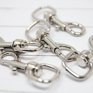 May include: Five silver metal swivel snap hooks. The hooks are all closed and are lying on a white wooden surface.