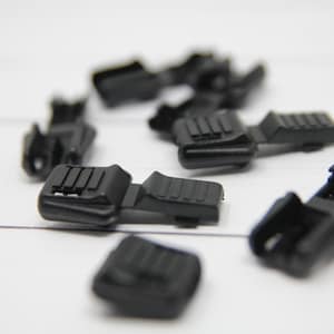 May include: A group of eight black plastic zipper pulls scattered on a white surface. The zipper pulls have a rectangular shape with a raised textured surface.