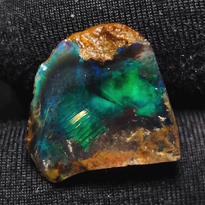 May include: A colourful opal gemstone, displaying a play of colour. The stone has a mix of green, blue, and gold hues, with a rough, textured edge. The opal's surface reflects light, creating a shimmering effect.