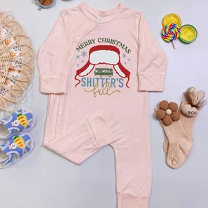 May include: A light pink baby onesie with long sleeves and attached feet. The onesie features a graphic with the text "MERRY CHRISTMAS" and "SHITTER'S full" in red, green, and gold. The onesie is surrounded by baby accessories.