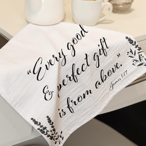 Op de afbeelding: Witte theedoek met de tekst "Every good & perfect gift is from above" in zwart schrift. De handdoek heeft een bloemenontwerp en staat naast een witte theepot, kop en een fruitmand.