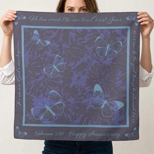 May include: A square scarf with a dark gray and purple floral pattern and light blue butterflies. The scarf has text that reads "He has created to nur love Christ Jesus," "For we are God's masterpiece," and "Ephesians 2:10 Happy Anniversary."