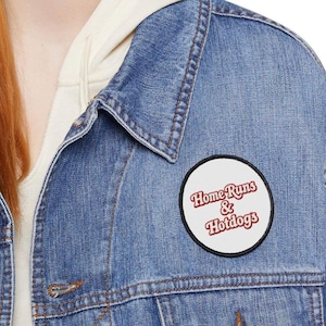 Peut inclure: Veste en jean bleu clair avec un écusson rond blanc orné de l'inscription "Home Runs & Hotdogs" en lettres rouges. Le patch est bordé de noir. Un sweat à capuche clair est visible en dessous.