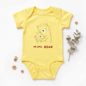May include: Yellow baby onesie with a cartoon bear graphic. The graphic includes a mother bear hugging a baby bear, with the word "LOVE" and hearts above them. The text "MIMI BEAR" is printed below the bears.