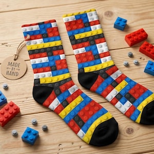 May include: A pair of colorful socks with a Lego brick design, featuring red, blue, yellow, black, and white blocks. The socks have black heels and toes. The tag says "Made by Hand". Several Lego bricks and studs are scattered around the socks.