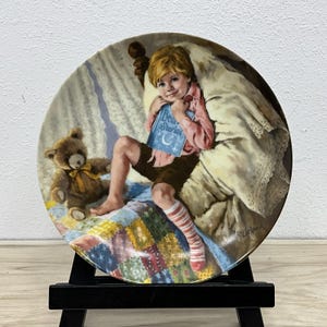 May include: Decorative plate featuring a young child sitting on a bed, holding a book titled "Bear Stories". A teddy bear sits nearby. The plate has a colorful quilt design and is displayed on a black easel.