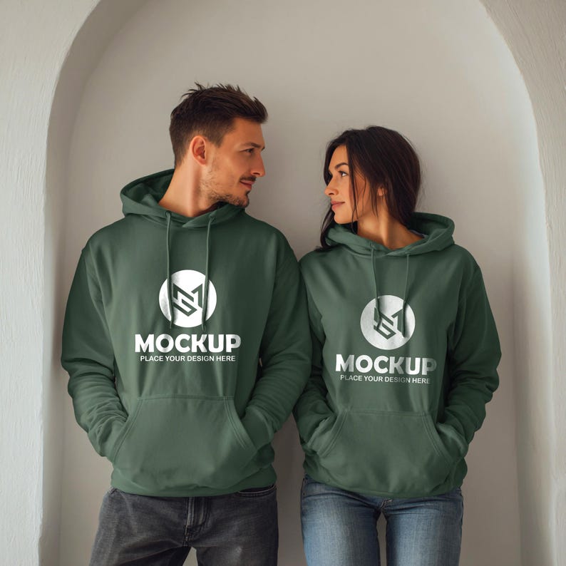 May include: Two people wearing matching dark green hoodies with a white logo and the text "MOCKUP PLACE YOUR DESIGN HERE". The hoodies have a front pocket and a hood. The people are standing in front of a white wall.