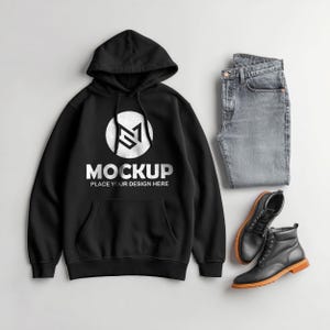May include: Black hoodie with a white logo and the word "MOCKUP" printed on the front. The hoodie is paired with gray jeans and black leather boots with brown soles. The outfit is laid out on a light gray surface.