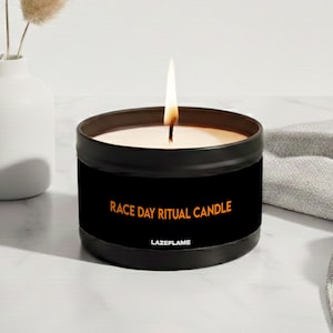 May include: A black metal candle tin with a lit wick, featuring the orange text "RACE DAY RITUAL CANDLE" and "LAZEFLAME" on the front label. The candle is positioned next to a white vase containing dried beige grass plumes.