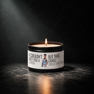May include: A black metal tin candle with a white label featuring a photograph of a race car driver and the text: I COULDN'T GET YOU A LANDO BUT THIS CANDLE SMELLS LIKE HIM. The candle has a single lit wick with a visible flame.