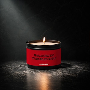 May include: A black metal candle tin with a red label featuring the text FERRARI STRATEGY STRESS RELIEF CANDLE and LAZEFLAME. The candle has a single lit wick with a visible flame, and the wax inside the container is off-white.