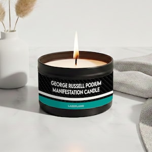 May include: A black metal candle tin with a lit wick, featuring a label that reads GEORGE RUSSELL PODIUM MANIFESTATION CANDLE and LAZEFLAME, accented with a white stripe and a teal band against a black carbon fiber pattern background.