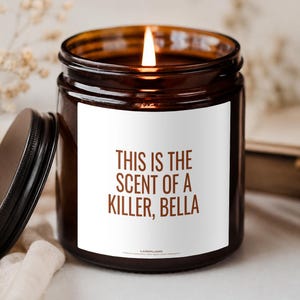 Könnte beinhalten: Eine braune Glas-Duftkerze mit brennendem Docht und einem weißen Etikett, das den braunen Text "THIS IS THE SCENT OF A KILLER, BELLA" sowie den Markennamen "LAZEFLAME" unten zeigt. Ein schwarzer Metalldeckel liegt neben der Kerze.