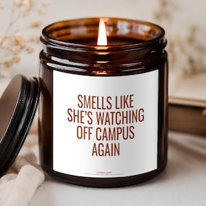 Könnte beinhalten: Eine braune Glasgefäßkerze mit brennendem Docht und einem weißen Etikett mit dem braunen Text: SMELLS LIKE SHE'S WATCHING OFF CAMPUS AGAIN. Die Kerze hat einen passenden braunen Deckel, der daneben liegt, und das Wachs leuchtet durch die Flamme.