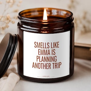 Può includere: Una candela accesa in un barattolo di vetro ambrato scuro con un coperchio nero posizionato di lato. L'etichetta bianca riporta il testo marrone: SMELLS LIKE EMMA IS PLANNING ANOTHER TRIP. La candela ha uno stoppino acceso al centro.