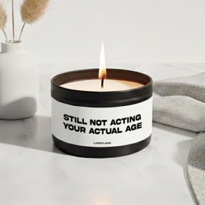 Può includere: Una candela in una scatola di metallo nero con un'etichetta bianca che riporta il testo nero "STILL NOT ACTING YOUR ACTUAL AGE" e "LAZEFLAME". La candela ha uno stoppino acceso con una fiamma visibile e la cera è di colore crema.