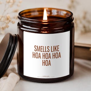 Könnte beinhalten: Eine brennende Kerze in einem dunkel bernsteinfarbenen Glasgefäß mit schwarzem Deckel. Das weiße Etikett trägt den braunen Text: SMELLS LIKE HOA HOA HOA HOA. Die Kerze hat einen einzelnen brennenden Docht und das Glasgefäß ist zylindrisch geformt.
