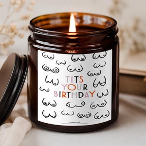 Könnte beinhalten: Eine brennende Kerze in einem braunen Glasgefäß mit schwarzem Deckel. Das weiße Etikett zeigt ein sich wiederholendes schwarzes Strichzeichnungsmuster von Brüsten und den bunten Text TITS YOUR BIRTHDAY. Der Markenname LAZEFLAME steht unten.