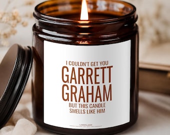 Può includere: Una candela in barattolo di vetro marrone con stoppino acceso e un'etichetta bianca con il testo marrone: I COULDN'T GET YOU GARRETT GRAHAM BUT THIS CANDLE SMELLS LIKE HIM. Il coperchio nero è appoggiato accanto su una superficie chiara.