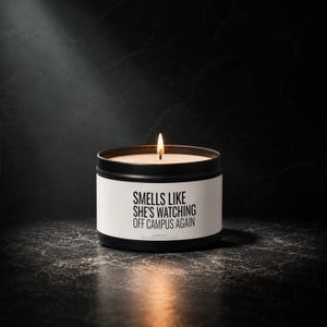 Op de afbeelding: Een zwarte metalen kaarsenblik met een wit etiket met de zwarte tekst "SMELLS LIKE SHE'S WATCHING OFF CAMPUS AGAIN". De kaars heeft een enkele brandende lont met een zichtbare vlam en de was in de houder heeft een crèmekleurige tint.