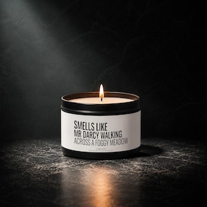 Könnte beinhalten: Eine schwarze Metalldose mit einer weißen Banderole, auf der in schwarzer Schrift steht: SMELLS LIKE MR DARCY WALKING ACROSS A FOGGY MEADOW. Die Kerze hat einen brennenden Docht mit einer sichtbaren Flamme und das Wachs ist cremefarben.