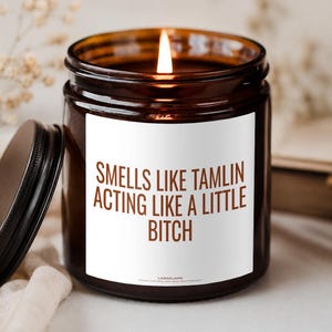 Könnte beinhalten: Eine braune Glas-Kerze mit brennendem Docht und einem weißen Etikett mit dem braunen Text: SMELLS LIKE TAMLIN ACTING LIKE A LITTLE BITCH. Die Kerze hat einen schwarzen Deckel, der daneben liegt, und das Wachs im Inneren ist dunkel gefärbt.